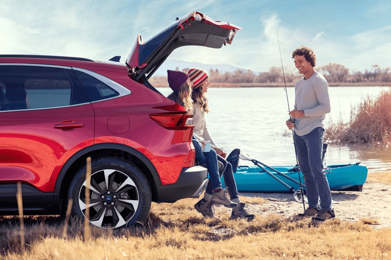 Family fishing by a red SUV near a lake with kayak in the background.